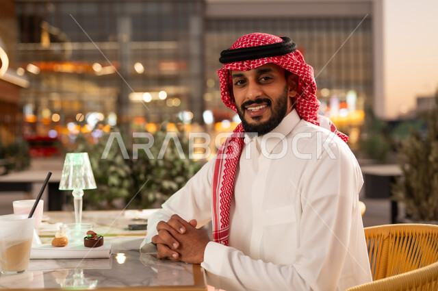 النظر إلى الكاميرا بإيماءات السعادة، الاستمتاع بتناول المشروب والحلا المفضل في الخارج، قضاء وقت ممتع في مطعم وكافيه راقي، شاب عربي خليجي سعودي يرتدي الثوب والشماغ يجلس في مقهى يقوم بشرب العصير المنعش