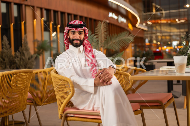 جلسة ترفيهية مسائية، الاستمتاع بتناول العصير المنعش في الخارج، قضاء وقت ممتع في مطعم وكافيه راقي، شاب عربي خليجي سعودي يرتدي الثوب والشماغ يجلس في مقهى ينظر إلى الكاميرا بإيماءات السعادة والسرور والثقة بالنفس