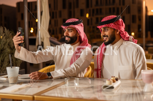 Evening youth outdoor party, taking a souvenir selfie using a mobile phone with happy and pleased gestures, having a cold and refreshing drink and dessert, entertainment and fun with friends, two Saudi Arabian Gulf men wearing traditional Saudi clothing sitting in a cafe