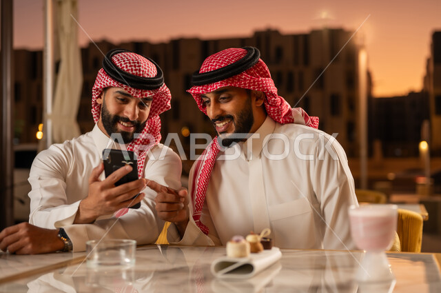 An outdoor evening youth party, watching entertainment programs and clips via mobile phone, having fun and entertainment with friends, two Saudi Arabian Gulf men wearing traditional Saudi clothing sitting in a cafe, sharing a cold drink and dessert with expressions of happiness and pleasure