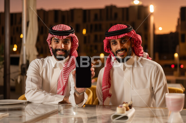 عرض شاشة سوداء فارغة عبر الهاتف المحمول، سهرة شبابية خارجية مسائية، التسلية والترفيه والمتعه مع الأصدقاء، رجلان عربيان خليجيان سعوديان يرتديان الزي التقليدي السعودي يجلسان في المقهى، النظر إلى الكاميرا بإيماءات السعادة والسرور