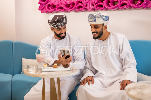 تصفح مواقع التواصل الاجتماعي، قضاء وقت ممتع مع الأصدقاء، رجلان عربيان خليجيان عمانيان يرتديان الدشداشة والعمامه يجلسان على الأريكة في المقهى يقومان بمشاهدة البرامج والمقاطع الترفيهية عبر الهاتف المحمول، المشاركة في تناول المشروب البارد والحلوى بتعابير السعادة