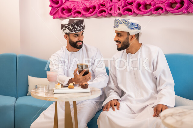 تصفح مواقع التواصل الاجتماعي، قضاء وقت ممتع مع الأصدقاء، رجلان عربيان خليجيان عمانيان يرتديان الدشداشة والعمامه يجلسان على الأريكة في المقهى يقومان بمشاهدة البرامج والمقاطع الترفيهية عبر الهاتف المحمول، المشاركة في تناول المشروب البارد والحلوى بتعابير السعادة