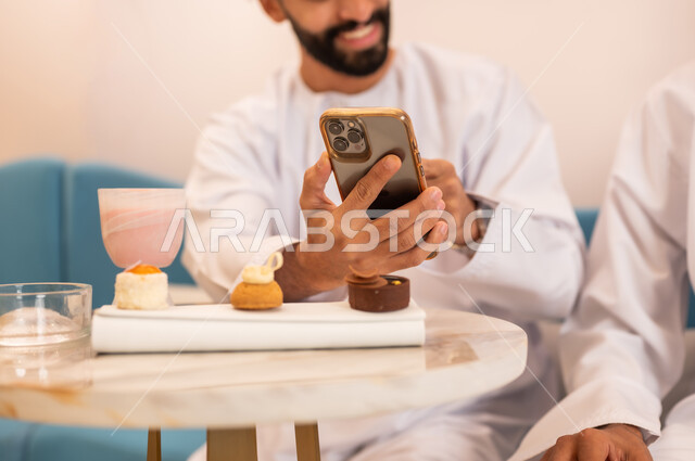 المشاركة في تناول المشروب البارد والحلوى، قضاء وقت ممتع مع الأصدقاء، صورة مقربة لرجل عربي خليجي عماني مبتسم يرتدي الدشداشه يجلس مع صديقه في المقهى ويتصفح مواقع الانترنت عبر الهاتف المحمول بإيماءات السعادة والسرور، استخدام جهاز تقني حديث ومتطور