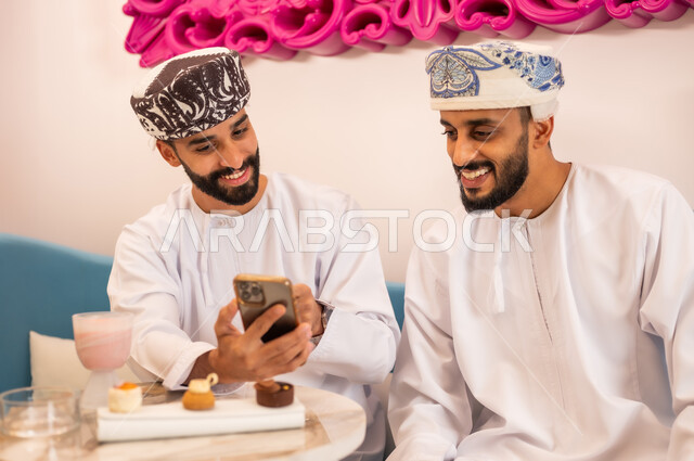 المشاركة في تناول المشروب البارد والحلوى بتعابير السعادة، تصفح مواقع التواصل الاجتماعي، قضاء وقت ممتع مع الأصدقاء، رجلان عربيان خليجيان عمانيان يرتديان الدشداشة والعمامه يجلسان على الأريكة في المقهى يقومان بمشاهدة البرامج والمقاطع الترفيهية عبر الهاتف المحمول