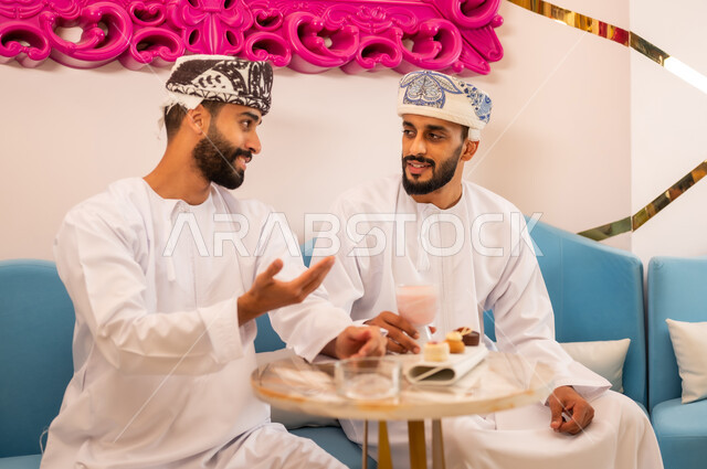 اجتماع الرفاق على تناول المشروب البارد والحلوى، جلسة وناسة في عطلة نهاية الاسبوع, ايماءات السعادة والسرور، قضاء وقت ممتع برفقة الأصدقاء، صديقان عربيان خليجيان عمانيان يرتديان الدشداشة والعمامه يجلسان على الأريكة في مقهى راقي يقومان بتبادل أطراف الحديث