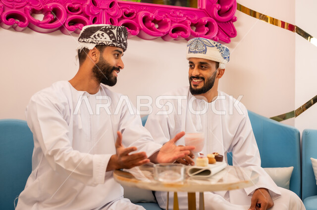 اجتماع الرفاق على تناول المشروب البارد والحلوى، جلسة وناسة في عطلة نهاية الاسبوع, ايماءات السعادة والسرور، قضاء وقت ممتع برفقة الأصدقاء، صديقان عربيان خليجيان عمانيان يرتديان الدشداشة والعمامه يجلسان على الأريكة في مقهى راقي يقومان بتبادل أطراف الحديث