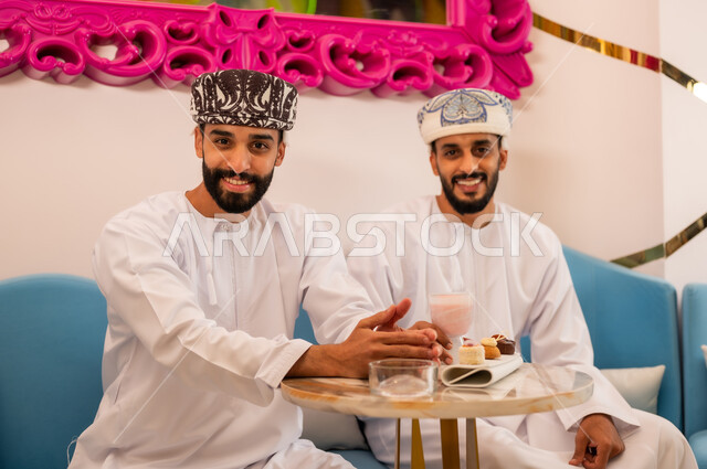 النظر إلى الكاميرا يايماءات السعادة والسرور، قضاء وقت ممتع برفقة الأصدقاء، جلسة وناسة في عطلة نهاية الاسبوع, صديقان عربيان خليجيان عمانيان يرتديان الدشداشة والعمامه يجلسان على الأريكة في مقهى راقي يقومان بتناول المشروب البارد والحلوى
