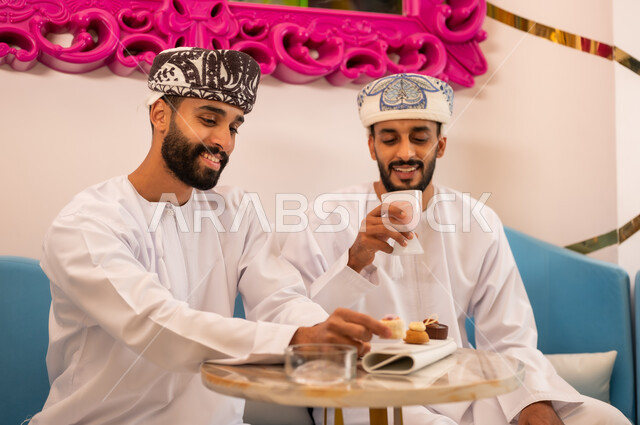 قضاء وقت ممتع برفقة الأصدقاء، جلسة وناسة في عطلة نهاية الاسبوع, ايماءات السعادة والسرور، صديقان عربيان خليجيان عمانيان يرتديان الدشداشة والعمامه يجلسان على الأريكة في مقهى راقي يقومان بتناول المشروب البارد والحلوى