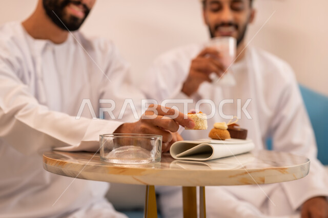 تعابير السعادة والسرور، قضاء وقت ممتع برفقة الأصدقاء، جلسة وناسة في عطلة نهاية الاسبوع, صورة مقربة لصديقين عربيين خليجيين عمانيين يرتديان الدشداشة والعمامه يجلسان على الأريكة في مقهى راقي يقومان بتناول المشروب البارد والحلوى