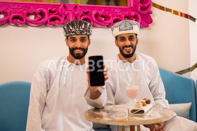 تناول المشروب البارد والحلوى في الخارج مع الأصدقاء، الإعجاب بتطبيقات الجهاز التقني الحديث، رجل عربي خليجي عماني مبتسم يرتدي الدشداشه والعمامه يجلس مع صديقه في المقهى يقوم بعرض شاشة سوداء فارغة عبر الهاتف المحمول، النظر إلى الكاميرا بإيماءات السرور والسعادة