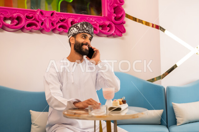 التواصل مع الأهل والأصدقاء، تناول المشروب البارد والحلوى في الخارج، استخدام الجهاز التقني الحديث، رجل عربي خليجي عماني مبتسم يرتدي الدشداشه والعمامه يجلس على الاريكه في المقهى يقوم بإجراء مكالمة هاتفية عبر الهاتف المحمول بإيماءات السرور والسعادة