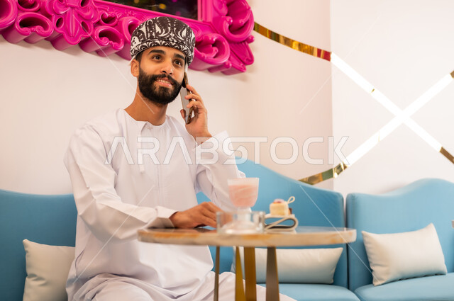 قضاء وقت ممتع بتناول المشروب البارد والحلوى في الخارج، التواصل مع الأهل والأصدقاء، استخدام الجهاز التقني الحديث، رجل عربي خليجي عماني مبتسم يرتدي الدشداشه والعمامه يجلس على الاريكه في المقهى يقوم بإجراء مكالمة هاتفية عبر الهاتف المحمول