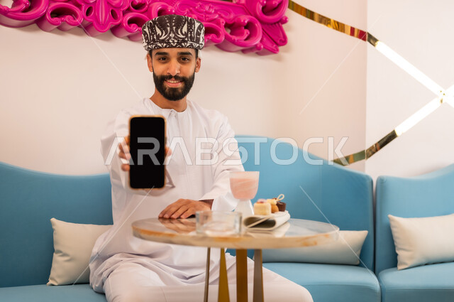 الإعجاب بتطبيقات الجهاز التقني الحديث، رجل عربي خليجي عماني مبتسم يرتدي الدشداشه والعمامه يجلس على الأريكة في المقهى يقوم بعرض شاشة سوداء فارغة عبر الهاتف المحمول، النظر إلى الكاميرا بإيماءات السرور والسعادة، تناول المشروب البارد والحلوى في الخارج