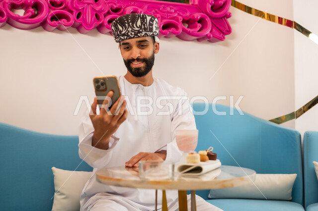 قضاء وقت ممتع ومسلي في الكافيه، رجل عربي خليجي عماني مبتسم يرتدي الدشداشه والعمامه يجلس على الأريكة في المقهى ويتصفح مواقع الانترنت عبر الهاتف المحمول بإيماءات السعادة والسرور، استخدام جهاز تقني حديث ومتطور