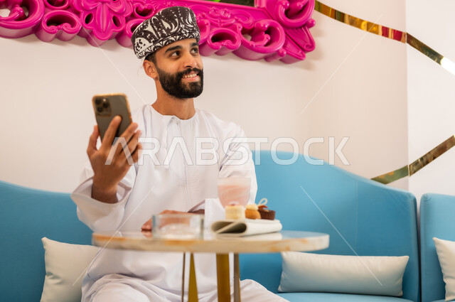 استخدام جهاز تقني حديث ومتطور، قضاء وقت ممتع ومسلي في الكافيه، رجل عربي خليجي عماني مبتسم يرتدي الدشداشه والعمامه يجلس على الأريكة في المقهى ويسمك بيده الهاتف المحمول، النظر إلى شيء ما بإيماءات السعادة والسرور