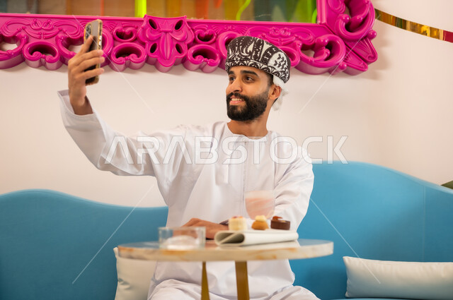 Gestures of happiness and pleasure, having a good time having a cold drink and dessert outside, using a modern technical device, a smiling Omani Gulf Arab man wearing a dishdasha and turban sitting on the sofa in a cafe taking a souvenir selfie with his mobile phone