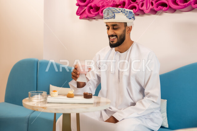 الاستمتاع بتناول المشروب والحلا المفضل في الخارج، قضاء وقت ممتع في مطعم وكافيه راقي في سلطنة عمان، شاب عربي خليجي عماني يرتدي الدشداشة والعمامة يجلس على الأريكة في مقهى يقوم بشرب السموذي المنعش بإيماءات السعادة