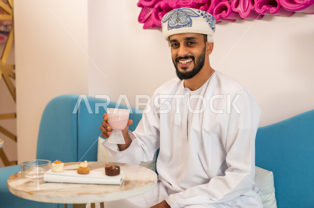 النظر إلى الكاميرا بإيماءات السعادة، الاستمتاع بتناول المشروب والحلا المفضل في الخارج، قضاء وقت ممتع في مطعم وكافيه راقي، شاب عربي خليجي عماني يرتدي الدشداشة والعمامة يجلس على الأريكة في مقهى يقوم بشرب السموذي المنعش