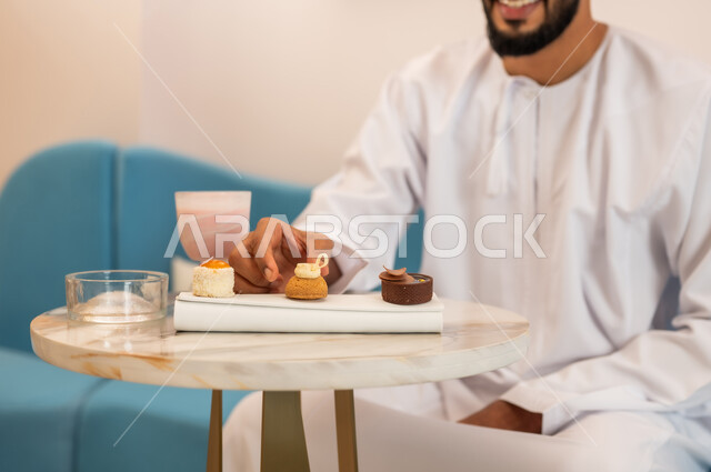 الاستمتاع بتناول المشروب والحلا المفضل، قضاء وقت ممتع في مطعم وكافيه راقي، صورة مقربة لطبق حلوى و لشاب عربي خليجي عماني يرتدي الدشداشة والعمامة يجلس على الأريكة في مقهى يقوم بأكل الحلوى