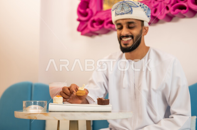 التسلية والترفيه في الخارج، إيماءات السعادة والسرور، الاستمتاع بتناول المشروب والحلا المفضل، قضاء وقت ممتع في مطعم وكافيه راقي، صورة مقربة لشاب عربي خليجي عماني يرتدي الدشداشة والعمامة يجلس على الأريكة في مقهى يقوم بأكل الحلوى