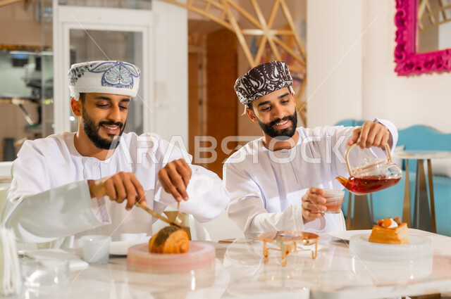 تعارف وصداقة وبناء علاقات اجتماعية، مقهى ومطعم راقي، جلسة وناسة يوم الإجازة, مشاركة المشروب الساخن والحلوى، صورة مقربة لرجلين عربيين خليجيين عمانيين يرتديان الدشداشه والعمامه يجلسان في كوفي شوب يتناولان الشاي والمخبوزات بإيماءات السعادة والسرور