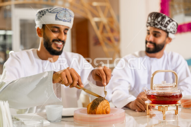 جلسة وناسة يوم الإجازة, مشاركة المشروب الساخن والحلوى، تعارف وصداقة وبناء علاقات اجتماعية، مقهى ومطعم راقي، صورة مقربة لرجلين عربيين خليجيين عمانيين يرتديان الدشداشه والعمامه يجلسان في كوفي شوب يتناولان الشاي والمخبوزات بإيماءات السعادة والسرور