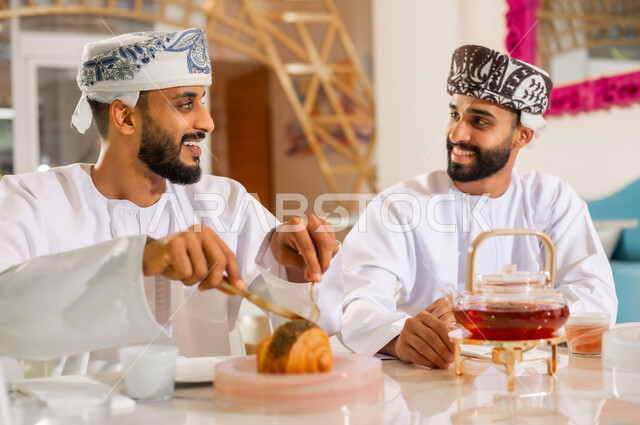 جلسة وناسة يوم الإجازة, مشاركة المشروب الساخن والحلوى، تعارف وصداقة وبناء علاقات اجتماعية، مقهى ومطعم راقي، صورة مقربة لرجلين عربيين خليجيين عمانيين يرتديان الدشداشه والعمامه يجلسان في كوفي شوب يتناولان الشاي والمخبوزات بإيماءات السعادة والسرور