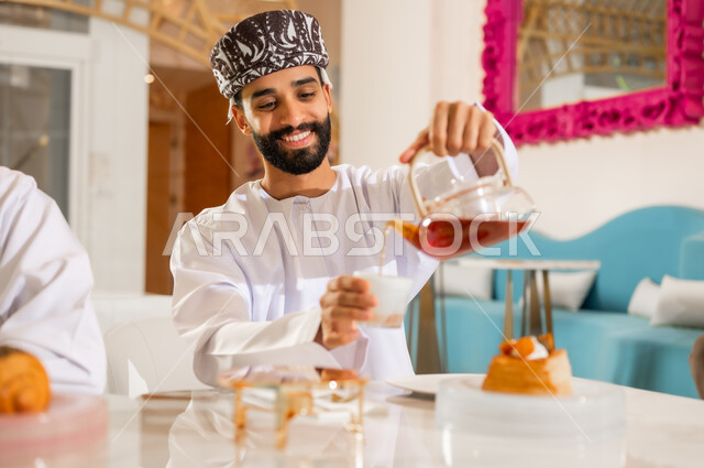 احتساء مشروب ساخن لذيذ, الاستمتاع بتناول المشروبات المفضلة, شاب عربي خليجي عماني يرتدي الدشداشة والعمامة يقوم بصب كوب الشاي بايماءات السعادة والسرور, التلذذ بمشاريب وحلوى الكافيهات والمطاعم الجاهزة
