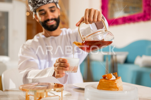 التلذذ بمشاريب وحلوى الكافيهات والمطاعم الجاهزة، احتساء مشروب ساخن لذيذ, الاستمتاع بتناول المشروبات المفضلة, شاب عربي خليجي عماني يرتدي الدشداشة والعمامة يقوم بصب كوب الشاي بايماءات السعادة والسرور