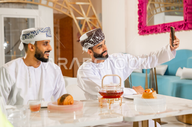 استخدام الوسائل والتقنيات الحديثة, مشاركة المشروب الساخن والمخبوزات، صديقان عربيان خليجيان عمانيان يرتديان الدشداشه والعمامة يجلسان في المقهى، قضاء وقت ممتع برفقة الأصدقاء خارج المنزل، التقاط الصور التذكارية عبر الهاتف المحمول بإيماءات السرور والسعادة