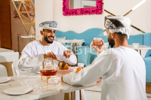 مشاركة المشروب الساخن والحلوى، تعارف وصداقة وبناء علاقات اجتماعية، جلسة وناسة في عطلة نهاية الاسبوع, مقهى ومطعم راقي، قضاء وقت ممتع في الخارج، صورة مقربة من الخلف لرجل عربي خليجي عماني يرتدي الدشداشه والعمامه يجلس مع صديقه في كوفي شوب  يتناولان الشاي