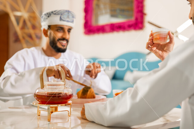 تعارف وصداقة وبناء علاقات اجتماعية، جلسة وناسة في عطلة نهاية الاسبوع, مقهى ومطعم راقي، مشاركة المشروب الساخن والحلوى، قضاء وقت ممتع في الخارج، رجل عربي خليجي عماني يرتدي الدشداشه والعمامه يجلس مع صديقه في كوفي شوب  يتناولان الشاي بإيماءات السعادة