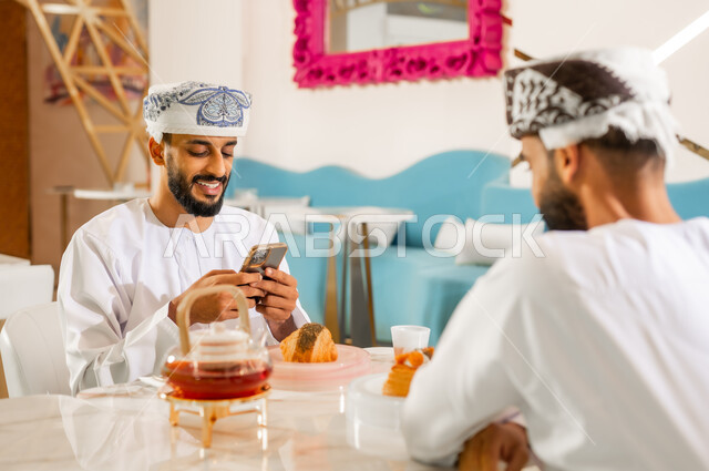 استخدام الوسائل والتقنيات الحديثة, مشاركة المشروب الساخن والمخبوزات، صديقان عربيان خليجيان عمانيان يرتديان الدشداشه والعمامة يجلسان في المقهى، قضاء وقت ممتع برفقة الأصدقاء خارج المنزل، رجل يقوم باستخدام الهاتف المحمول بإيماءات السرور والسعادة