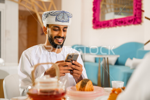 احتساء مشروب ساخن لذيذ، وسيلة تقنية حديثة ومتطورة، مطعم وكافيه راقي في سلطنة عمان، صورة مقربة لشاب عربي خليجي عماني مبتسم يرتدي الدشداشة والعمامة يجلس في مقهى يقوم باستخدام الهاتف المحمول، الاستمتاع بتناول المشروب والحلا المفضل في الخارج