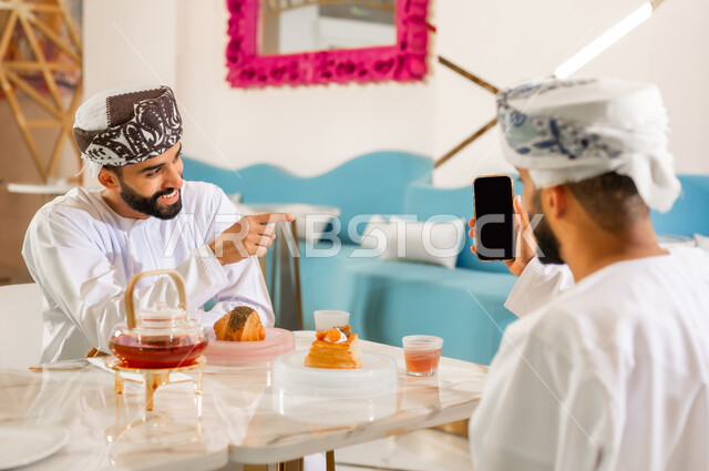 عرض شاشة سوداء فارغة، استخدام الوسائل والتقنيات الحديثة, مشاركة المشروب الساخن والمخبوزات، صديقان عربيان خليجيان عمانيان يرتديان الدشداشه والعمامة يجلسان في المقهى، قضاء وقت ممتع برفقة الأصدقاء خارج المنزل، الإشارة إلى شيء ما بإيماءات السرور