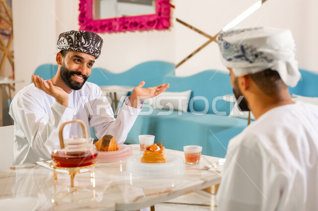 ايماءات عدم المعرفة، مشاركة المشروب الساخن والحلوى، تعارف وصداقة وبناء علاقات اجتماعية، جلسة وناسة في عطلة نهاية الاسبوع, مقهى ومطعم راقي، صورة مقربة من الخلف لرجل عربي خليجي عماني يرتدي الدشداشه والعمامه يجلس مع صديقه في كوفي شوب  يتناولان الشاي