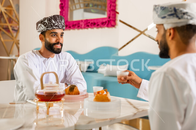 مقهى ومطعم راقي، مشاركة المشروب الساخن والحلوى، قضاء وقت ممتع في الخارج، صداقة وتعارف وبناء علاقات اجتماعية،  جلسة وناسة في عطلة نهاية الاسبوع, صورة مقربة من الخلف لرجل عربي خليجي عماني يرتدي الدشداشه والعمامه يجلس مع صديقه في كوفي شوب  يتناولان الشاي