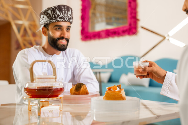 جلسة وناسة في عطلة نهاية الاسبوع, مقهى ومطعم راقي، مشاركة المشروب الساخن والحلوى، قضاء وقت ممتع في الخارج، صداقة وتعارف وبناء علاقات اجتماعية، رجل عربي خليجي عماني يرتدي الدشداشه والعمامه يجلس مع صديقه في كوفي شوب  يتناولان الشاي