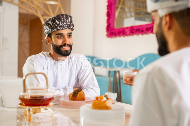 مقهى ومطعم راقي، مشاركة المشروب الساخن والحلوى، قضاء وقت ممتع في الخارج، صداقة وتعارف وبناء علاقات اجتماعية،  جلسة وناسة في عطلة نهاية الاسبوع, صورة مقربة من الخلف لرجل عربي خليجي عماني يرتدي الدشداشه والعمامه يجلس مع صديقه في كوفي شوب  يتناولان الشاي