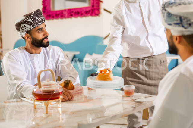 نادل يقوم بتقديم الحلوى، قضاء وقت ممتع في الخارج، صداقة وتعارف وبناء علاقات اجتماعية، الاهتمام والعناية بالزبائن، جلسة وناسة في عطلة نهاية الاسبوع, صديقان عربيان خليجيان عمانيان يرتديان الدشداشه والعمامه يجلسان في مقهى راقي، مشاركة المشروب الساخن
