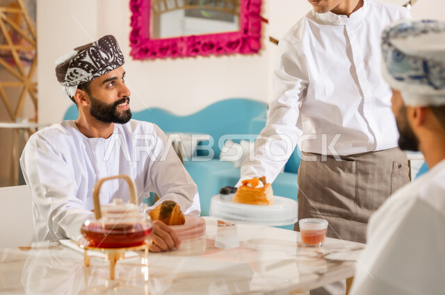 مشاركة المشروب الساخن، نادل يقوم بتقديم الحلوى، قضاء وقت ممتع في الخارج، صداقة وتعارف وبناء علاقات اجتماعية، الاهتمام والعناية بالزبائن، جلسة وناسة في عطلة نهاية الاسبوع, صديقان عربيان خليجيان عمانيان يرتديان الدشداشه والعمامه يجلسان في مقهى راقي