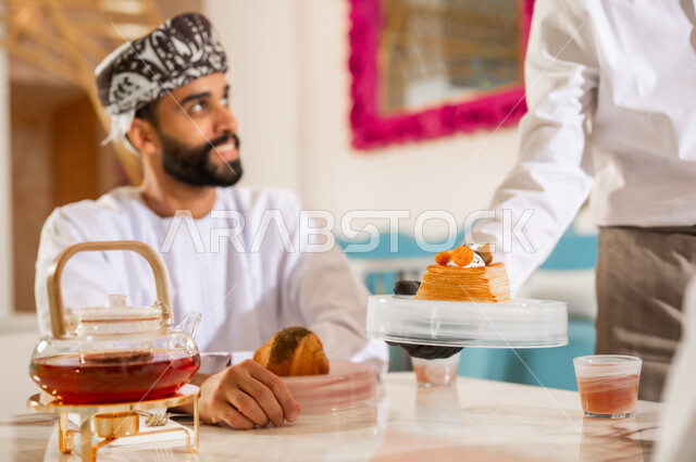 نادل يقوم بتقديم الحلوى، احتساء مشروب ساخن لذيذ، مطعم وكافيه راقي في سلطنة عمان، صورة مقربة لشاب عربي خليجي عماني يرتدي الدشداشة والعمامة يجلس في مقهى يقوم بشرب الشاي، الاستمتاع بتناول المشروب والحلا المفضل في الخارج، الاهتمام والعناية بالزبون