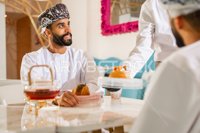 نادل يقوم بتقديم الحلوى، قضاء وقت ممتع في الخارج، صداقة وتعارف وبناء علاقات اجتماعية، الاهتمام والعناية بالزبائن، جلسة وناسة في عطلة نهاية الاسبوع, صديقان عربيان خليجيان عمانيان يرتديان الدشداشه والعمامه يجلسان في مقهى راقي، مشاركة المشروب الساخن