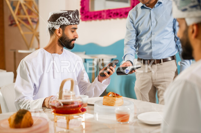 سهولة الدفع الالي في المقاهي والكافيهات في سلطنة عمان, رجل يقوم بدفع الحساب عبر الهاتف المحمول, استخدام الوسائل والتقنيات الحديثة في الدفع الالكتروني, مشاركة المشروب الساخن والمخبوزات، صورة مقربة لصديقين عربيين خليجيين عمانيين يرتديان الزي التقليدي يجلسان في المقهى