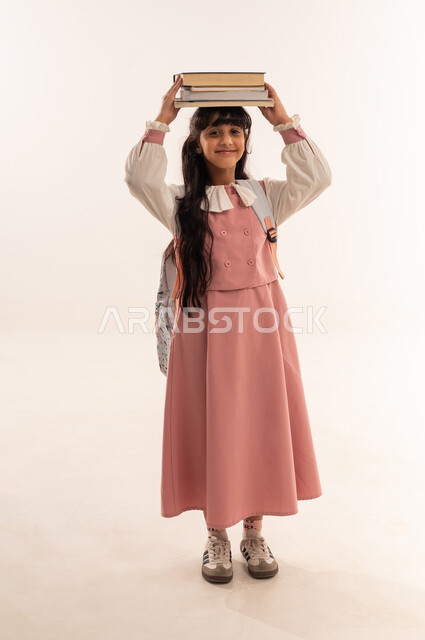 العودة الى المدارس، مفهوم الجد والاجتهاد للوصول إلى النجاح، المناهج العلمية التعليمية، مراجعة الدروس وحل الواجبات، بورتريه لطالبة عربيه خليجيه عمانية مبتسمه تحمل حقيبة الظهر المدرسية ، فتاه سعودية تحمل مجموعة من الكتب على رأسها، صورة لكامل الجسم، خلفية بيضاء