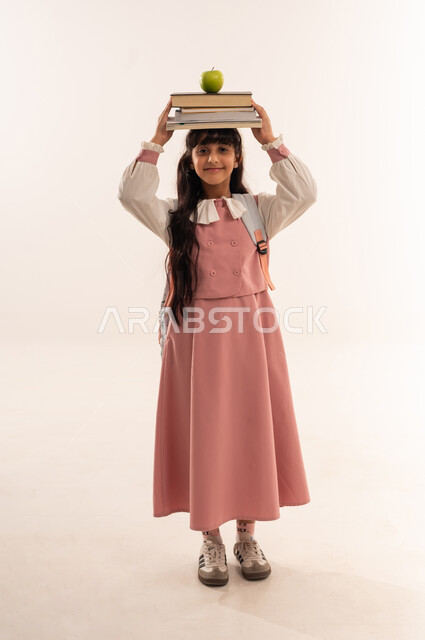 المناهج العلمية التعليمية، مفهوم الجد والاجتهاد للوصول إلى النجاح، العودة الى المدارس، بورتريه لطالبة عربيه خليجيه عمانية مبتسمه تحمل حقيبة الظهر المدرسية ، فتاه سعودية تحمل مجموعة من الكتب و تفاحه على رأسها، صورة لكامل الجسم، خلفية بيضاء