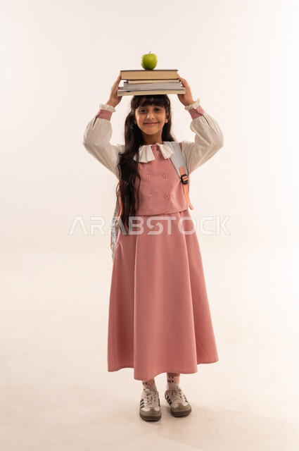 The concept of seriousness and diligence to achieve success. Back to school, educational scientific curricula, reviewing lessons and doing homework. Portrait of a smiling Arab Gulf Omani student carrying a school backpack. A Saudi girl carrying a group of books on her head. A full body image. White background.