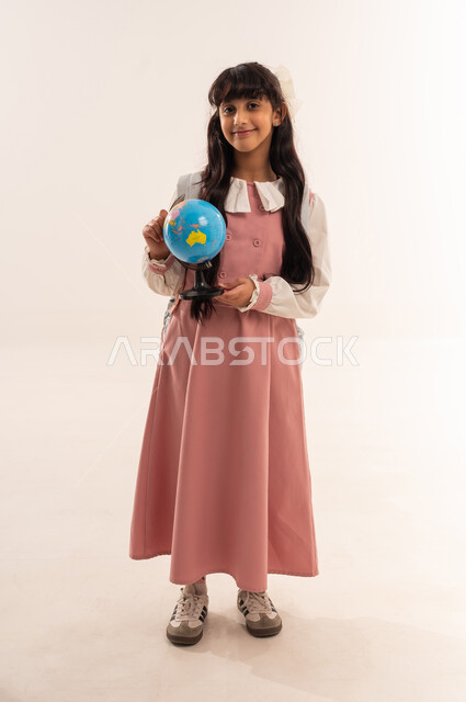 Back to school, preparing for the new school year, the concept of research and exploration around the world, a portrait of an Arab Gulf Omani student wearing a school uniform holding a globe, a Saudi girl looking with gestures of happiness and pleasure, full body, white background