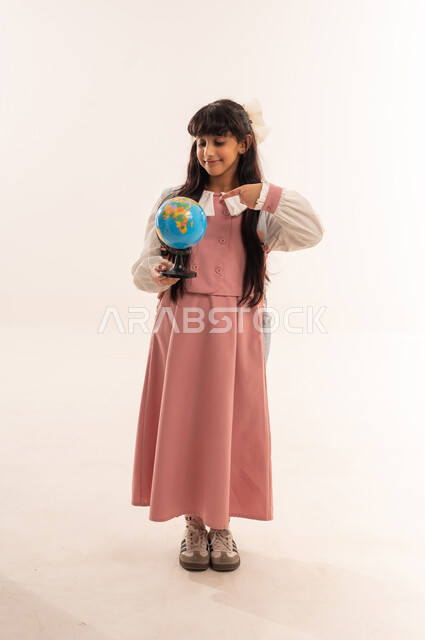 Preparing for the new school year, the concept of research and exploration around the world, back to school, portrait of an Arab Gulf Omani student wearing a school uniform holding a globe, a Saudi girl looking with gestures of happiness and pleasure, full body, white background